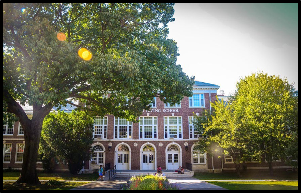 A large brick building with many windows and doors behind several green, leafy trees