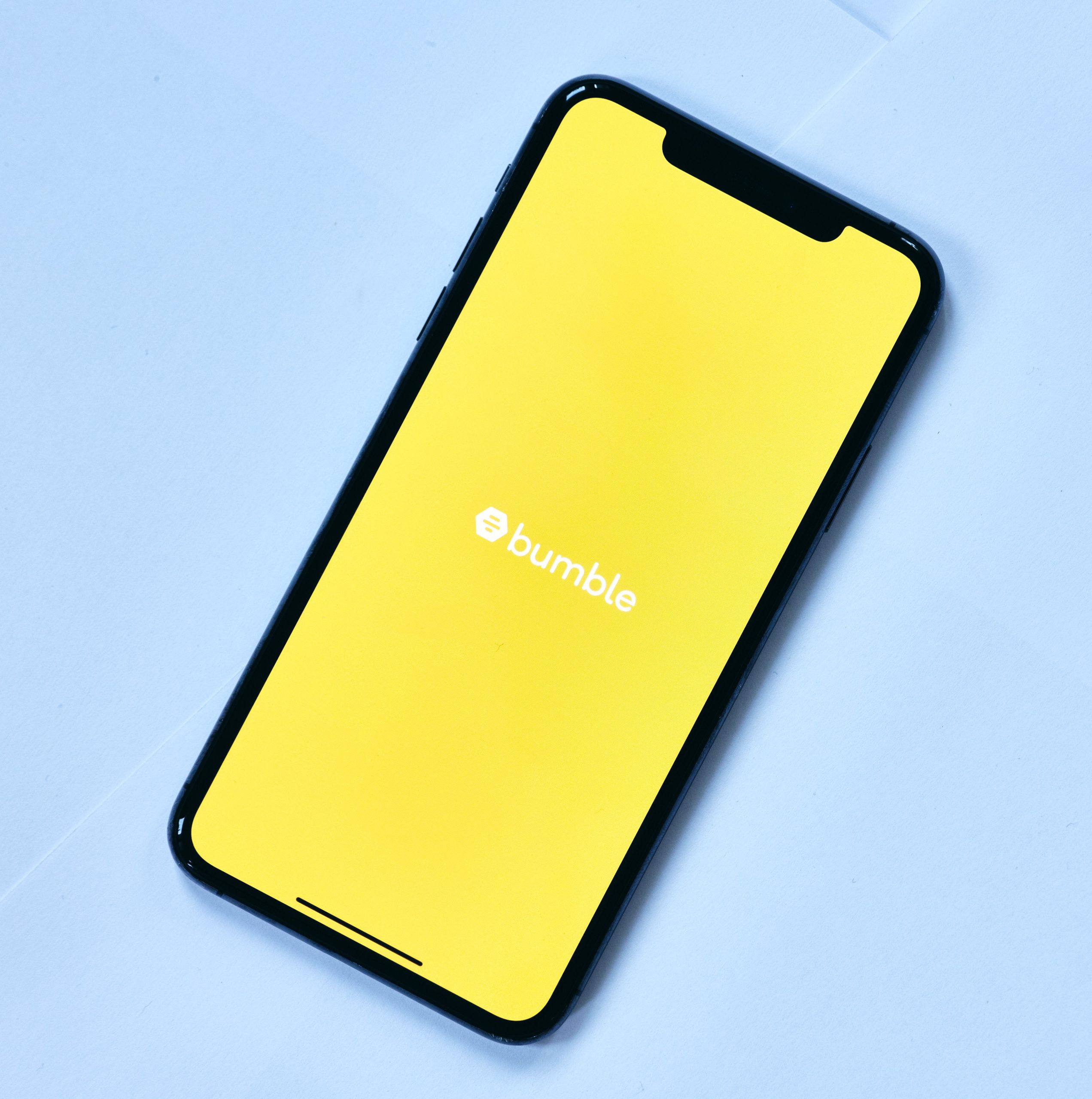 A Smartphone on a light blue background. The words "Bumble" are displayed in white on an all yellow background on the phone.