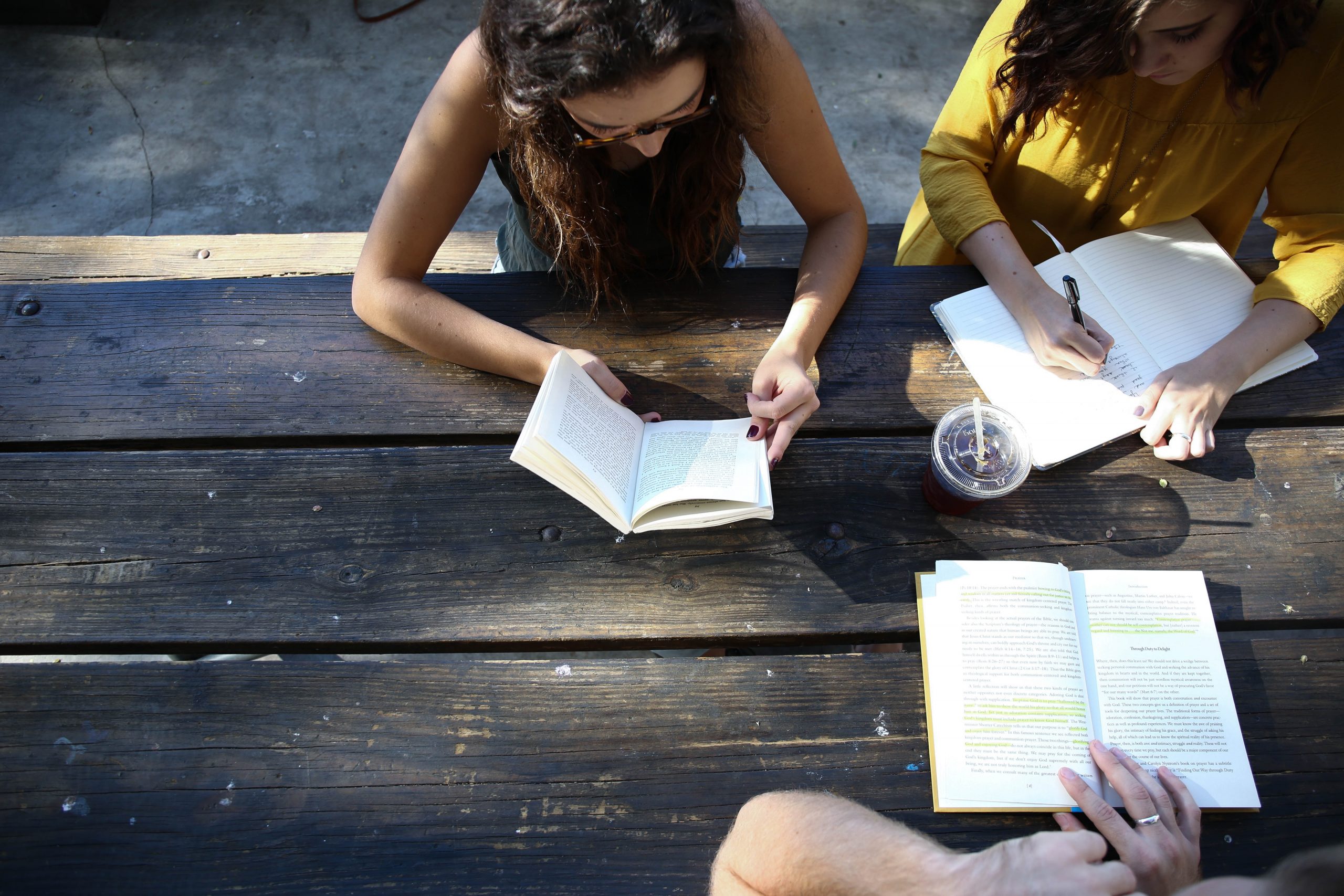 Three people look at books together while seated on an outdoor wooden table
