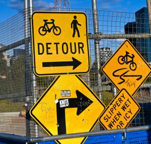 A bunch of signs on a pole. One says detour and another says slippery when wet